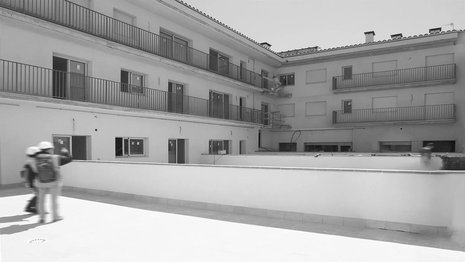 Fotografia en blanc i negre de les façanes interiors amb balcons correguts i dos tècnics amb casc a la coberta del soterrani.