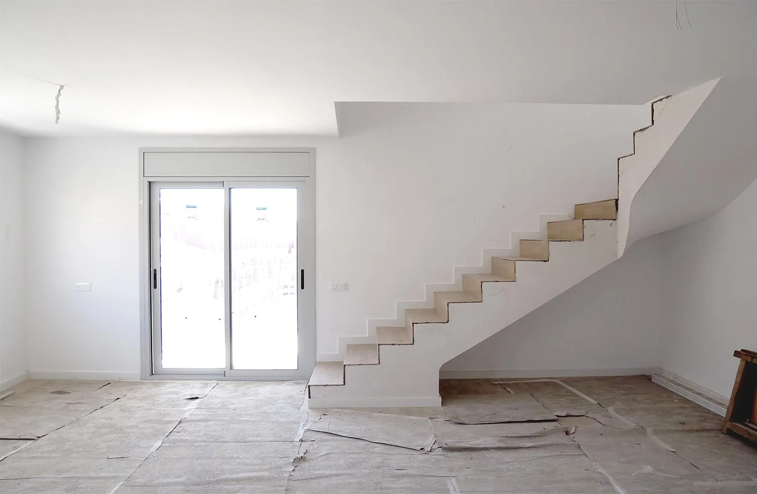 Interior d'un habitatge dúplex en obres amb una escala de formigó sense barana i una porta balconera, amb el terra protegit amb paper.