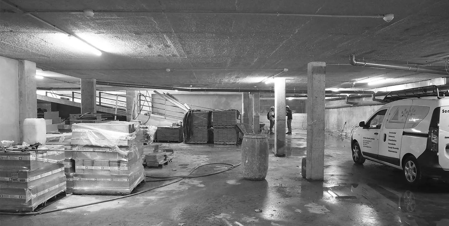 Fotografia en blanc i negre de l'aparcament soterrani en obres, amb materials apilats, pilars de formigó i una furgoneta.