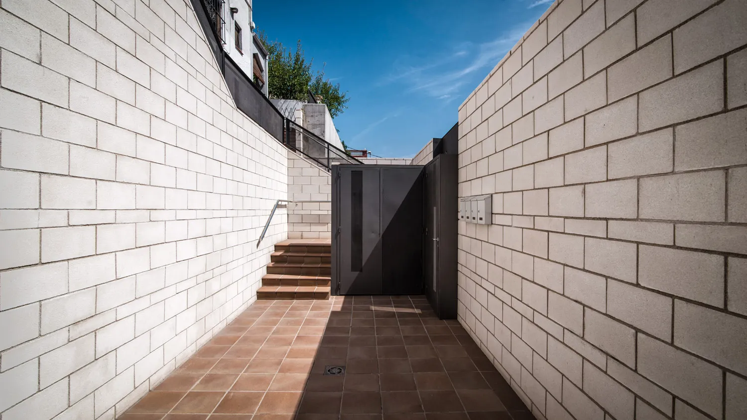 Escala exterior d'accés a la finca entre dos murs de bloc de formigó blanc, amb paviment ceràmic vermellós i una porta metàl·lica gris al fons.